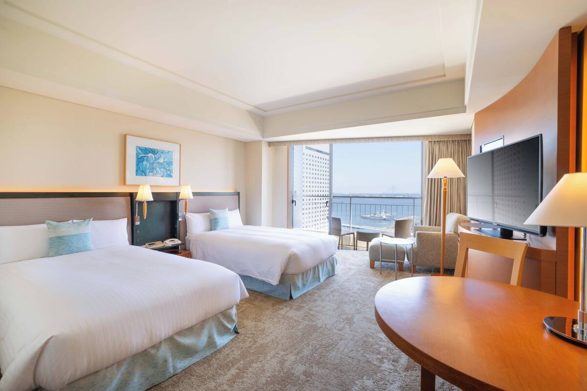 Habitación de hotel con dos camas, televisión, escritorio y balcón con vistas.