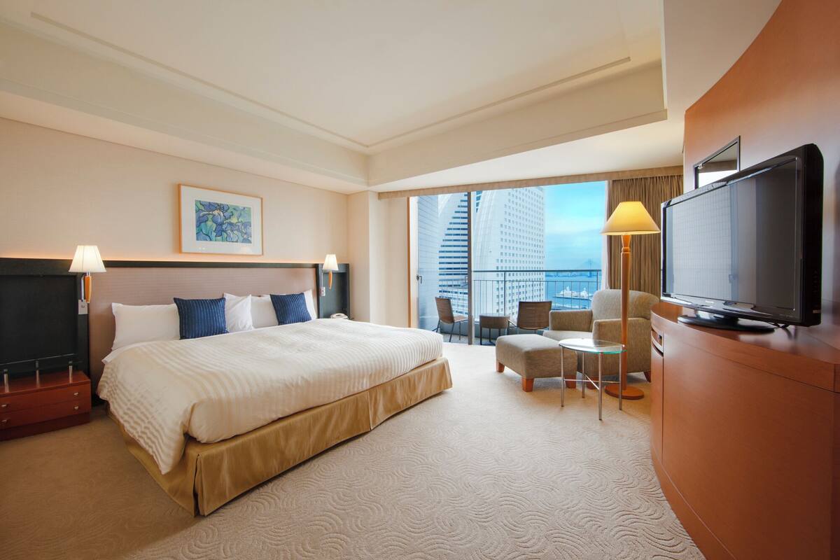 Habitación de hotel con una cama grande, televisión, escritorio con silla y vista a la ciudad por la ventana.