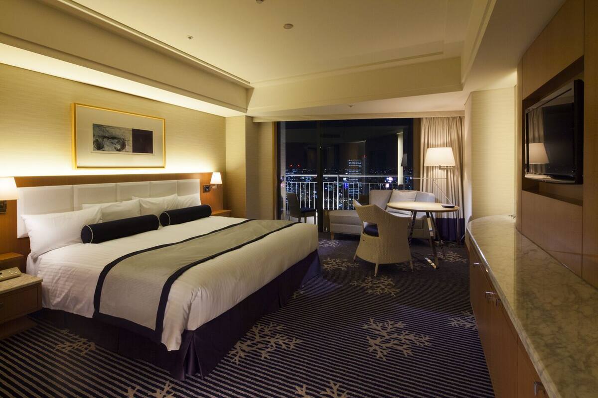 Habitación de hotel con una cama grande, un escritorio con silla, televisor y vista a la ciudad de noche.