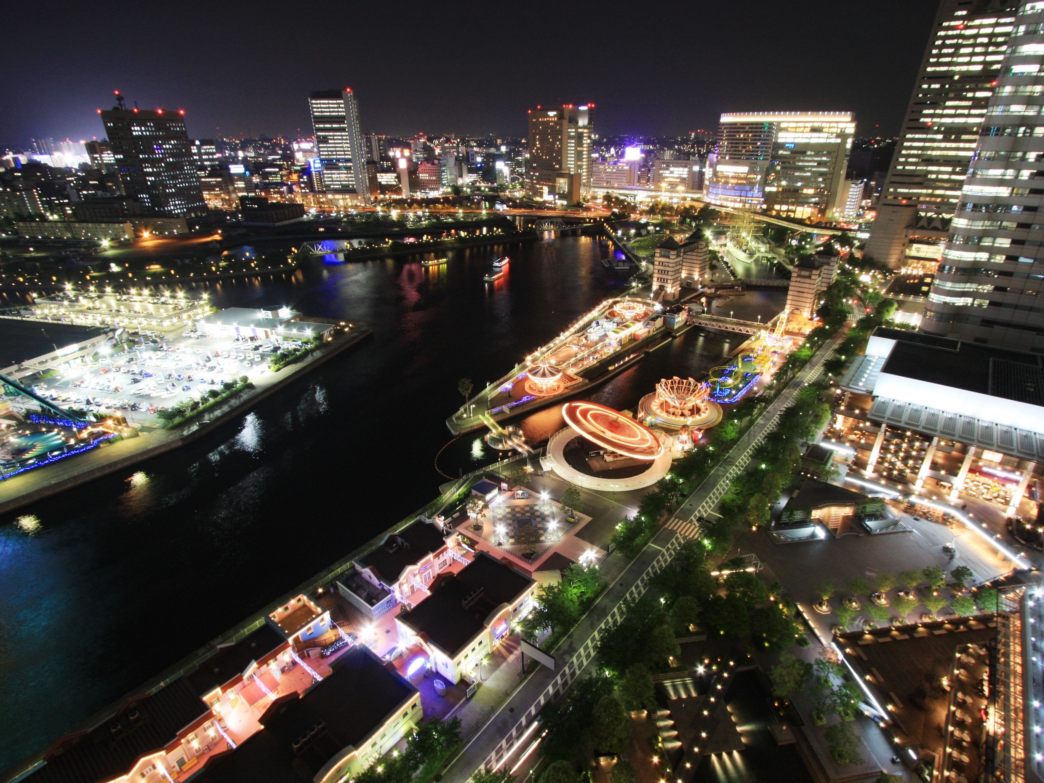 An aerial view of a cityscape at night with illuminated buildings, a river, and a waterfront promenade with lights and structures.