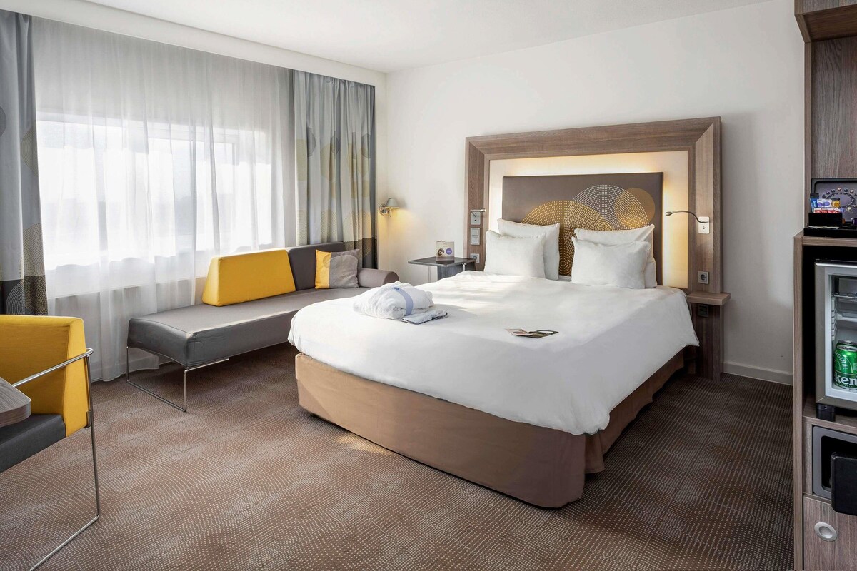 A hotel room with a large bed, a sofa, a desk, and a minibar.