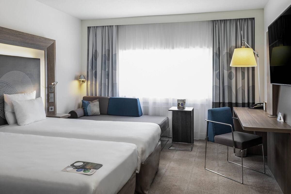 A hotel room with two beds, a desk, a chair, a lamp, and a TV.