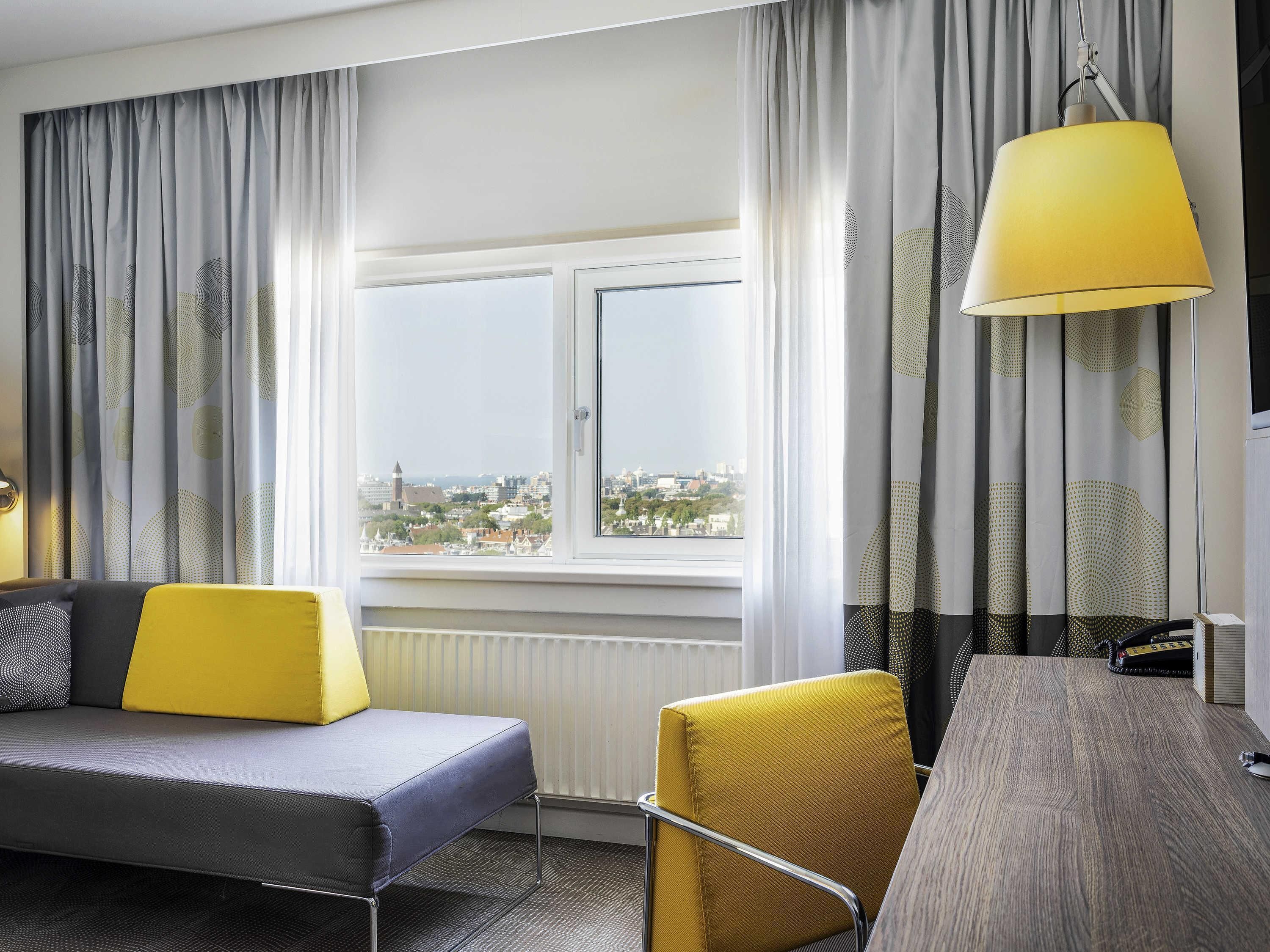 Ein Hotelzimmer mit Sofa, Sessel, Tisch, Lampe und einem Fenster mit Blick auf die Stadt.