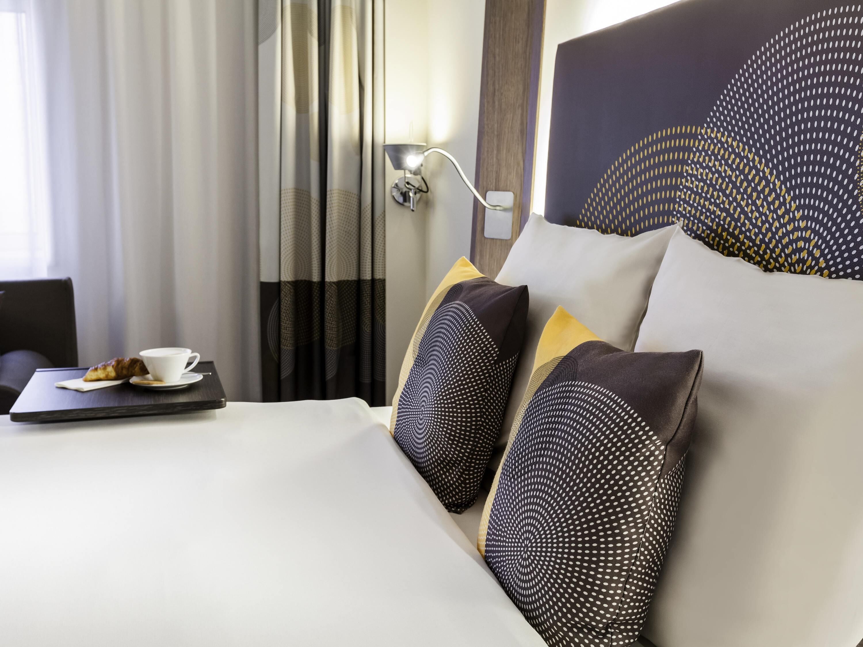 Ein Hotelzimmer mit einem aufgeräumten Bett, einem Nachttisch mit Tasse und Gebäck sowie einer Lampe.