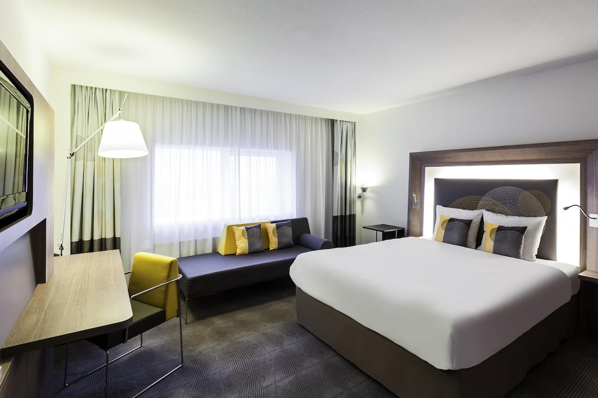 A hotel room with a large bed, a desk with a chair, a sofa, and a window with curtains.