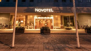 Um hotel com fachada de vidro moderna, entrada destacada com uma placa e dois mastro com bandeiras.