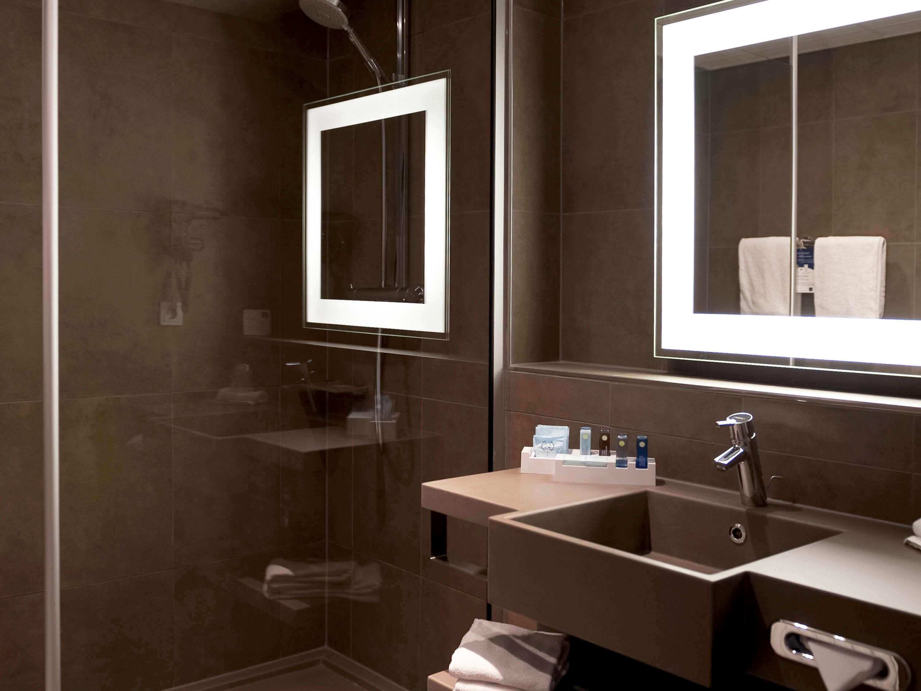 A modern bathroom with a glass shower enclosure, a sink with toiletries, and a large mirror.
