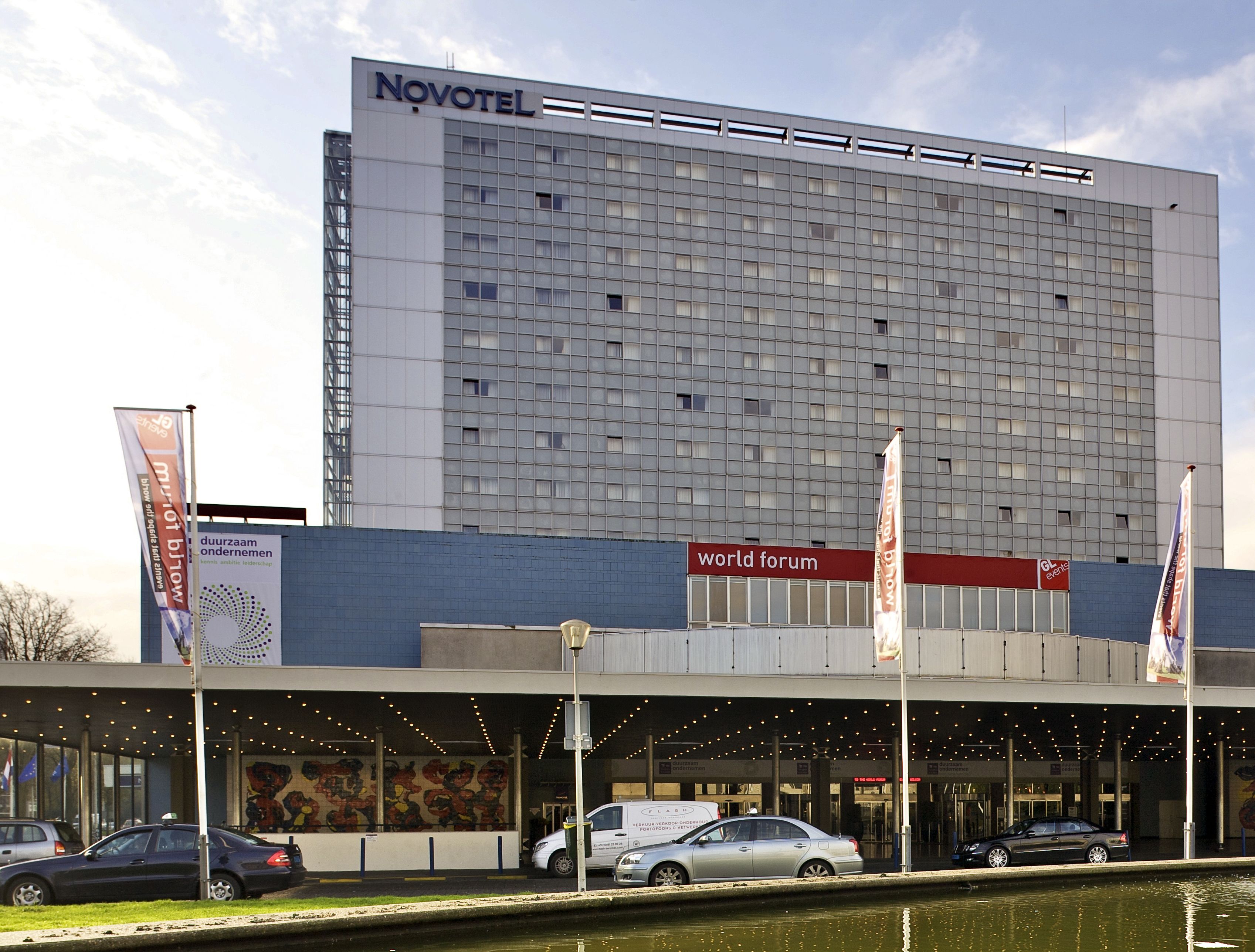 A hotel with a large, modern facade, a prominent 'Novotel' sign, and a 'World Forum' sign indicating a conference area.