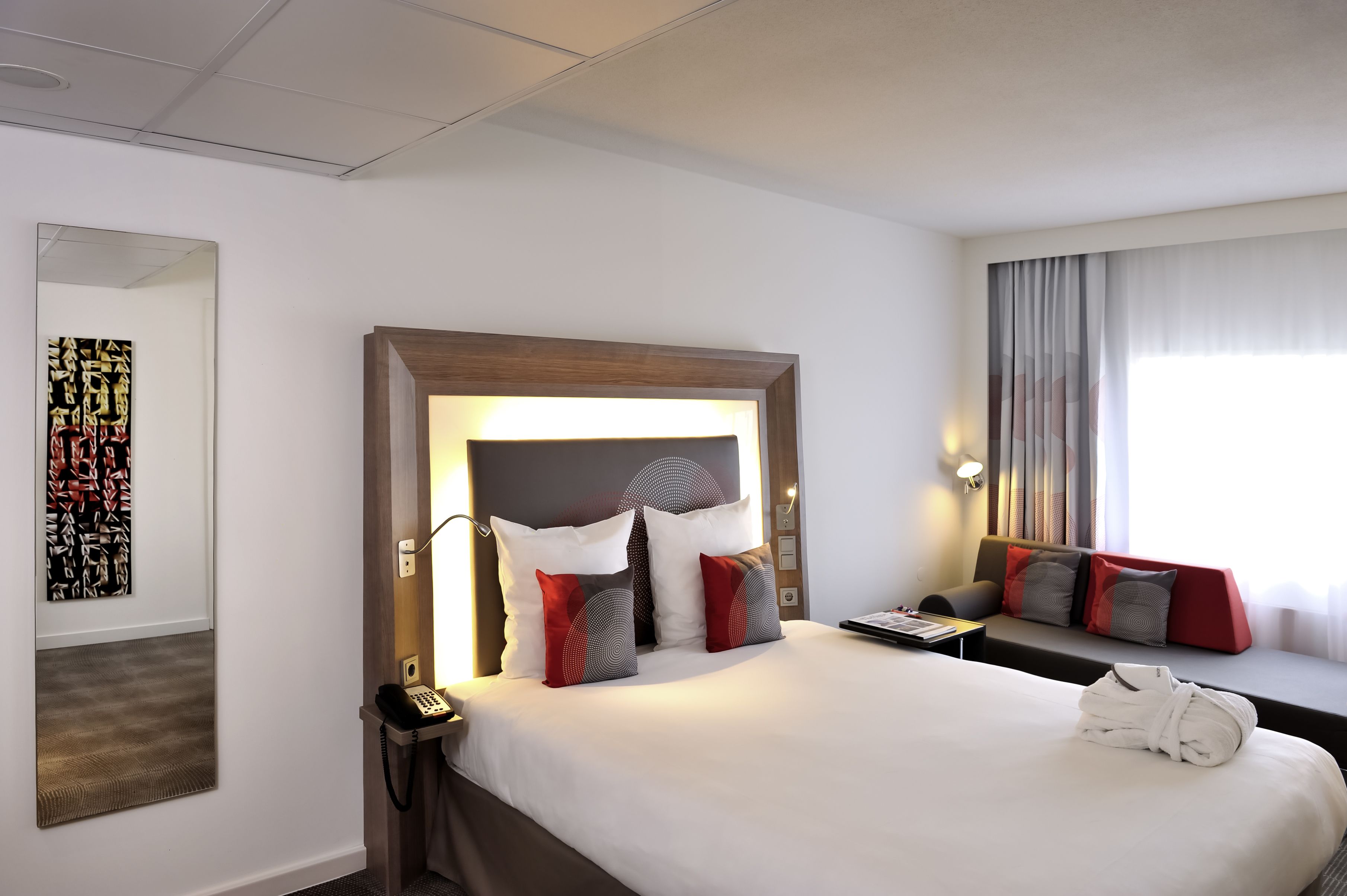 A hotel room with a large bed, a bedside table with a lamp, a telephone, and a window with curtains.