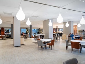 A modern dining area with tables and chairs, pendant lighting, and a view of the kitchen area. - Novotel Den Haag World Forum (The Hague)