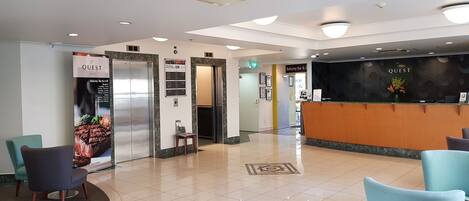 A hotel lobby with a reception desk, seating area, elevators, and a sign for the Balcony Bar & Grill.
