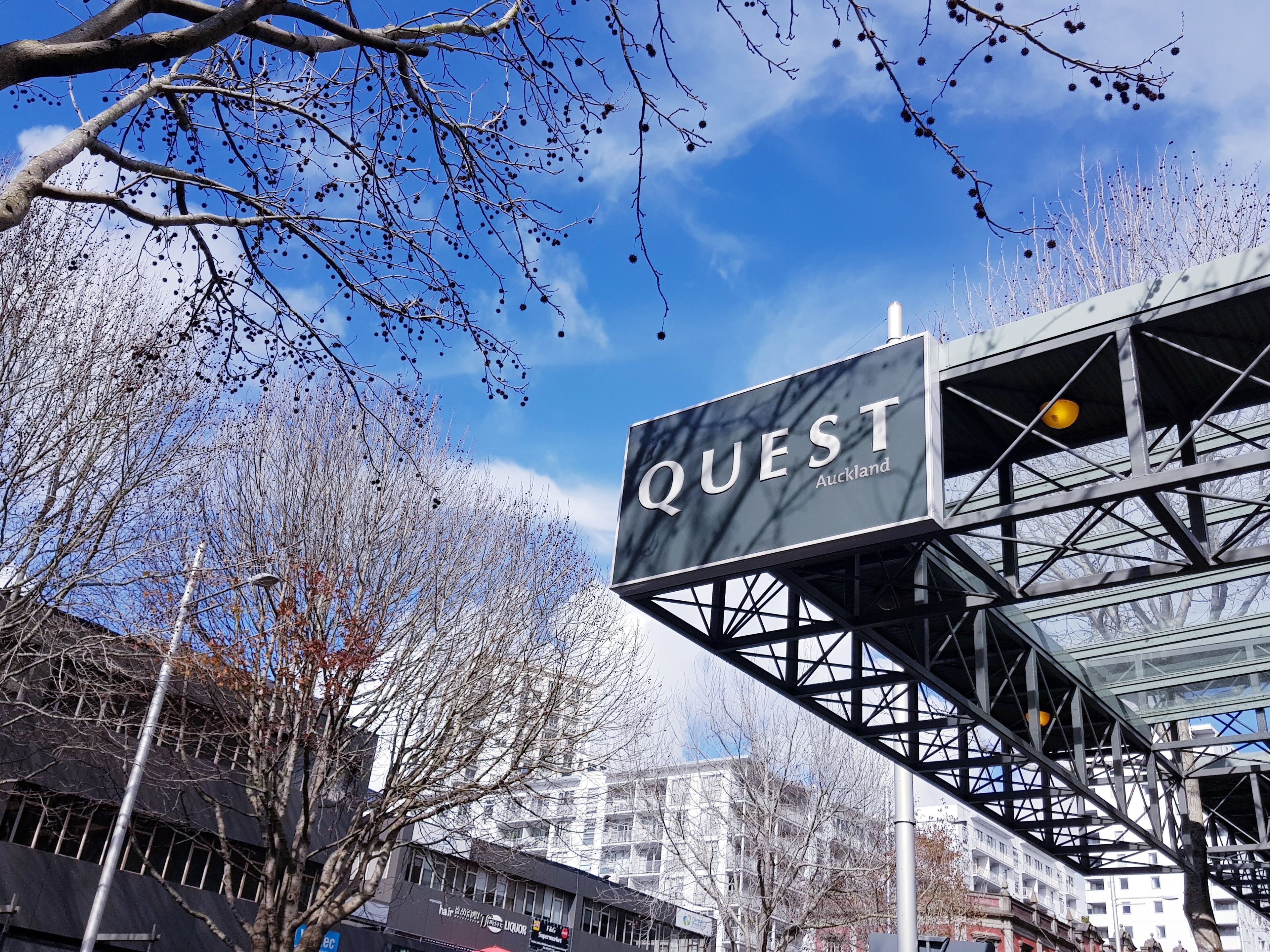 A sign for 'QUEST Auckland' with a modern metal structure and a clear blue sky in the background.