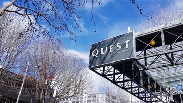 A sign for 'QUEST Auckland' with a modern metal structure and a clear blue sky in the background.