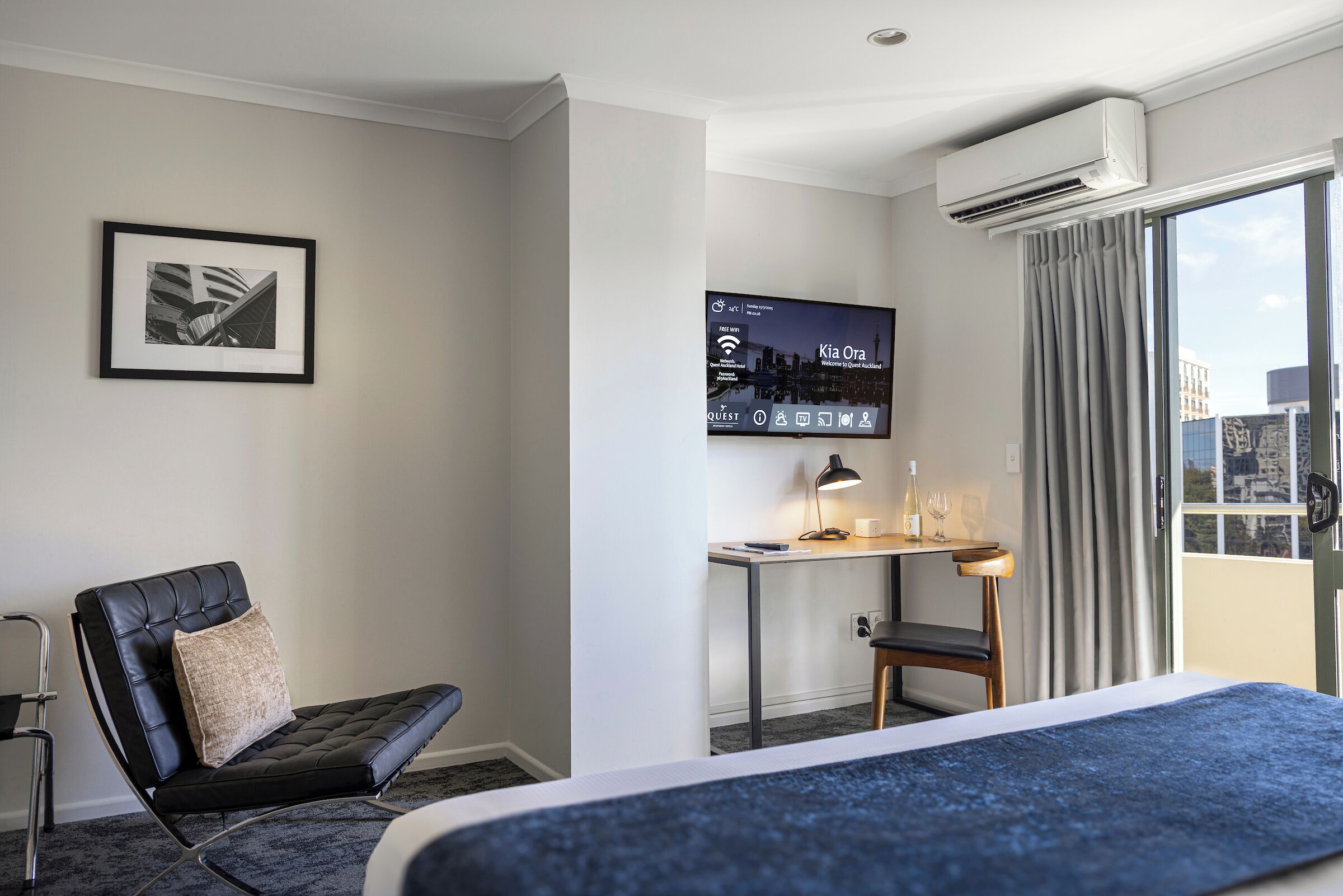 Ein Hotelzimmer mit einem Bett, einem Schreibtisch mit Lampe, einem Sessel, einem gerahmten Bild an der Wand und einem Fernseher, der eine Stadtansicht zeigt.