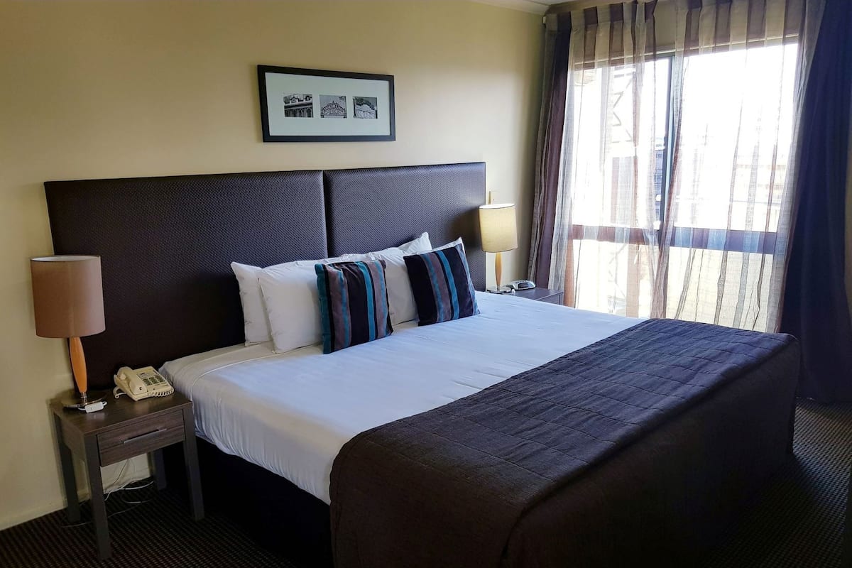 A hotel room with a large bed, a headboard, a bedside table with a lamp, a phone, and a framed picture on the wall.