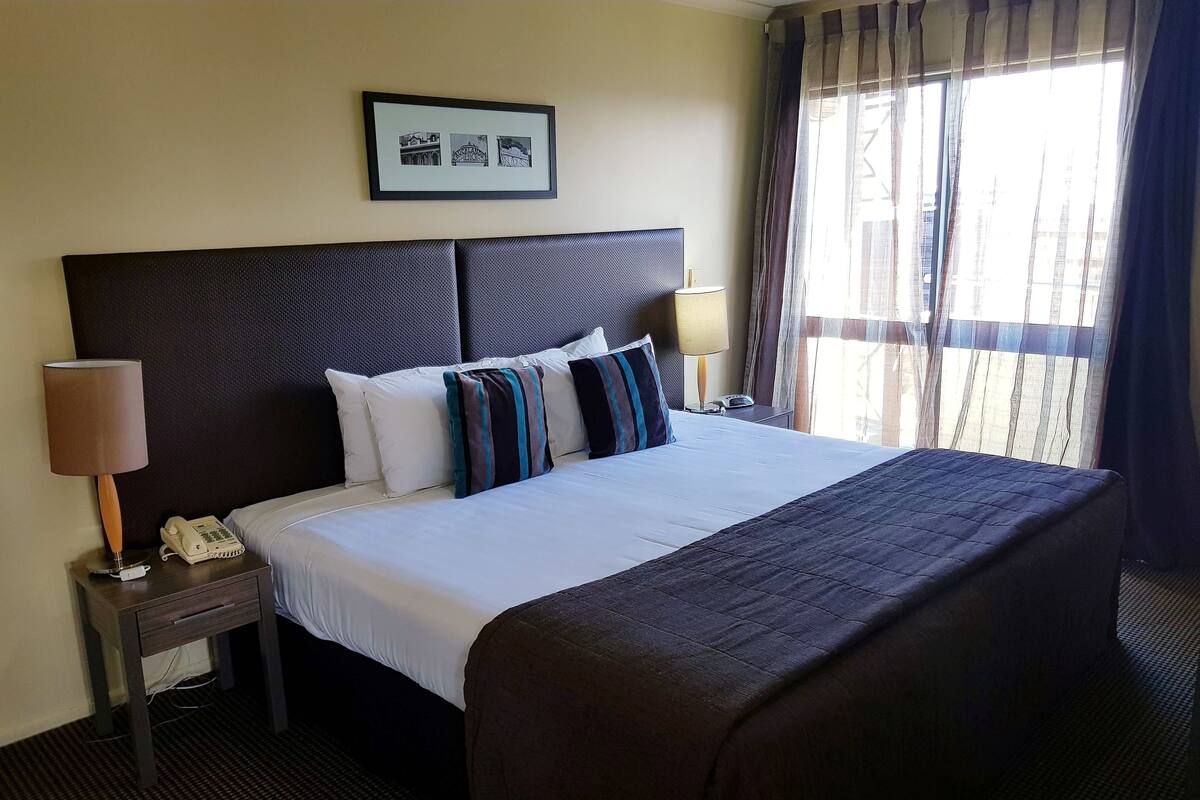 A hotel room with a large bed, a headboard, a bedside table with a lamp, a phone, and a framed picture on the wall.
