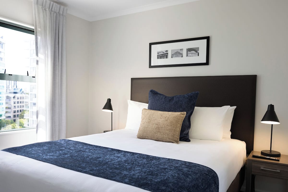 A neatly made bed with a dark blue blanket, two bedside lamps, a framed picture above the bed, and a window with curtains.