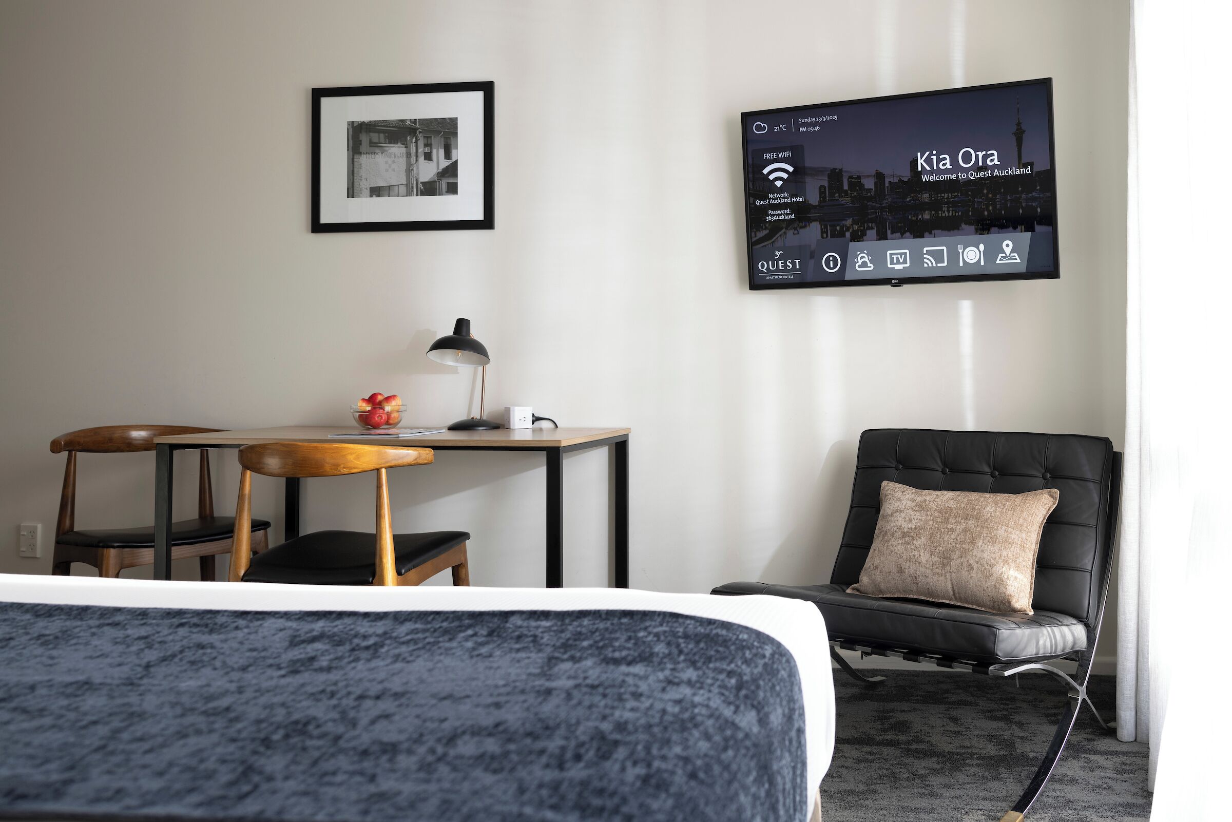 Habitación de hotel con cama, escritorio con silla, sillón de cuero negro, cuadro en la pared y televisor mostrando “Kia Ora Welcome to Quest Auckland”.