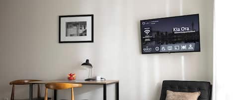 Habitación de hotel con cama, escritorio con silla, sillón de cuero negro, cuadro en la pared y televisor mostrando “Kia Ora Welcome to Quest Auckland”.