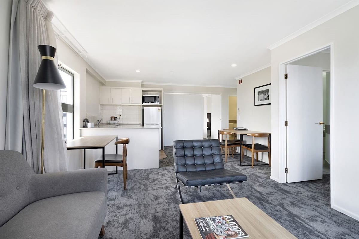 A modern living room with a grey sofa, a black leather chair, a wooden coffee table, and a dining area with a table and chairs.