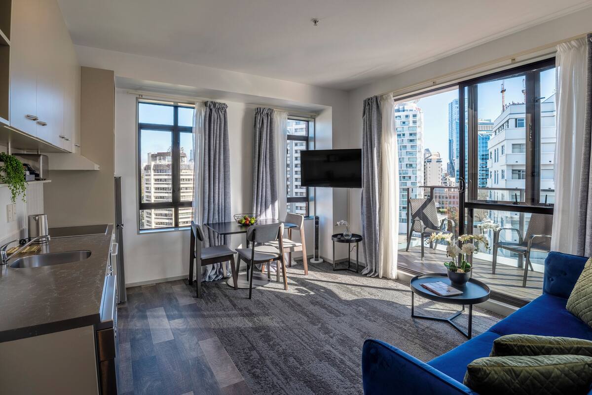 A modern apartment with a kitchen, dining area, and living room. It features large windows, a flat-screen TV, and a balcony with outdoor furniture.