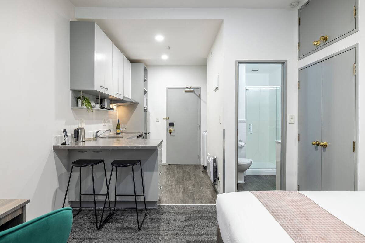 A compact hotel room with a kitchenette, a bed, a bathroom, and a small seating area.