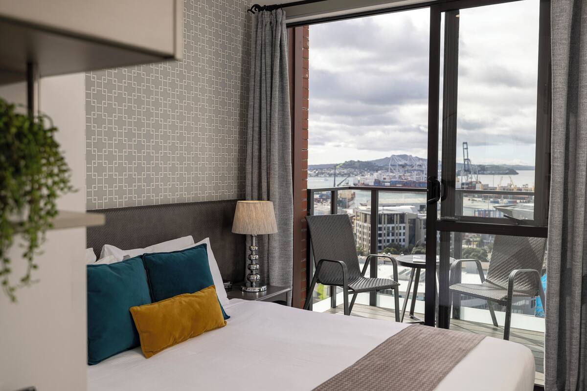 A hotel room with a large bed, a view of the city, and a balcony with chairs.