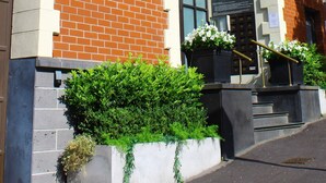 Un edificio de ladrillo con un letrero que dice "QUEST APARTMENTS". Hay escalones que conducen a la entrada, y macetas con plantas ubicadas en la base de los escalones.
