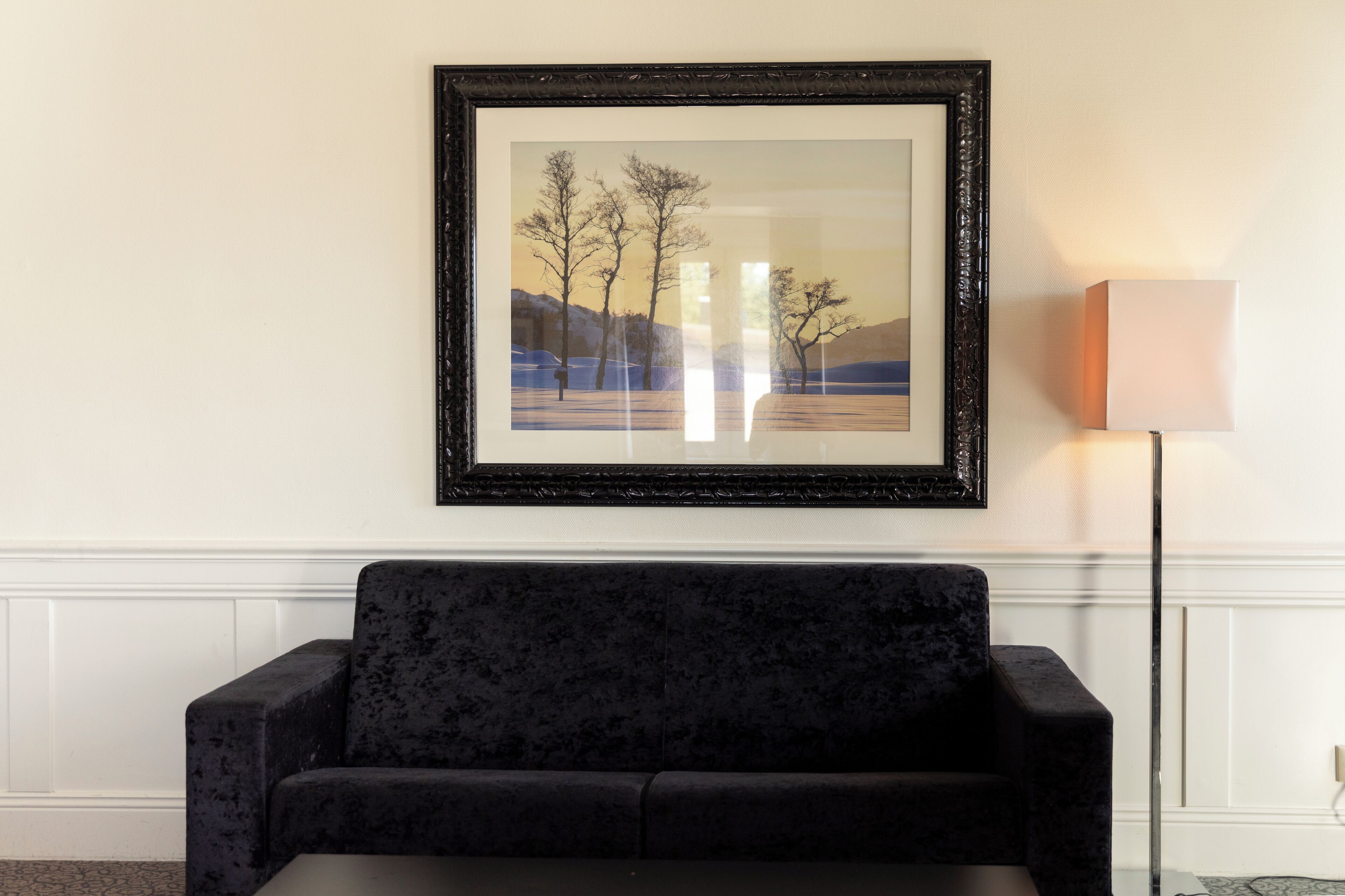 A framed landscape painting on a wall, a dark sofa, and a floor lamp.