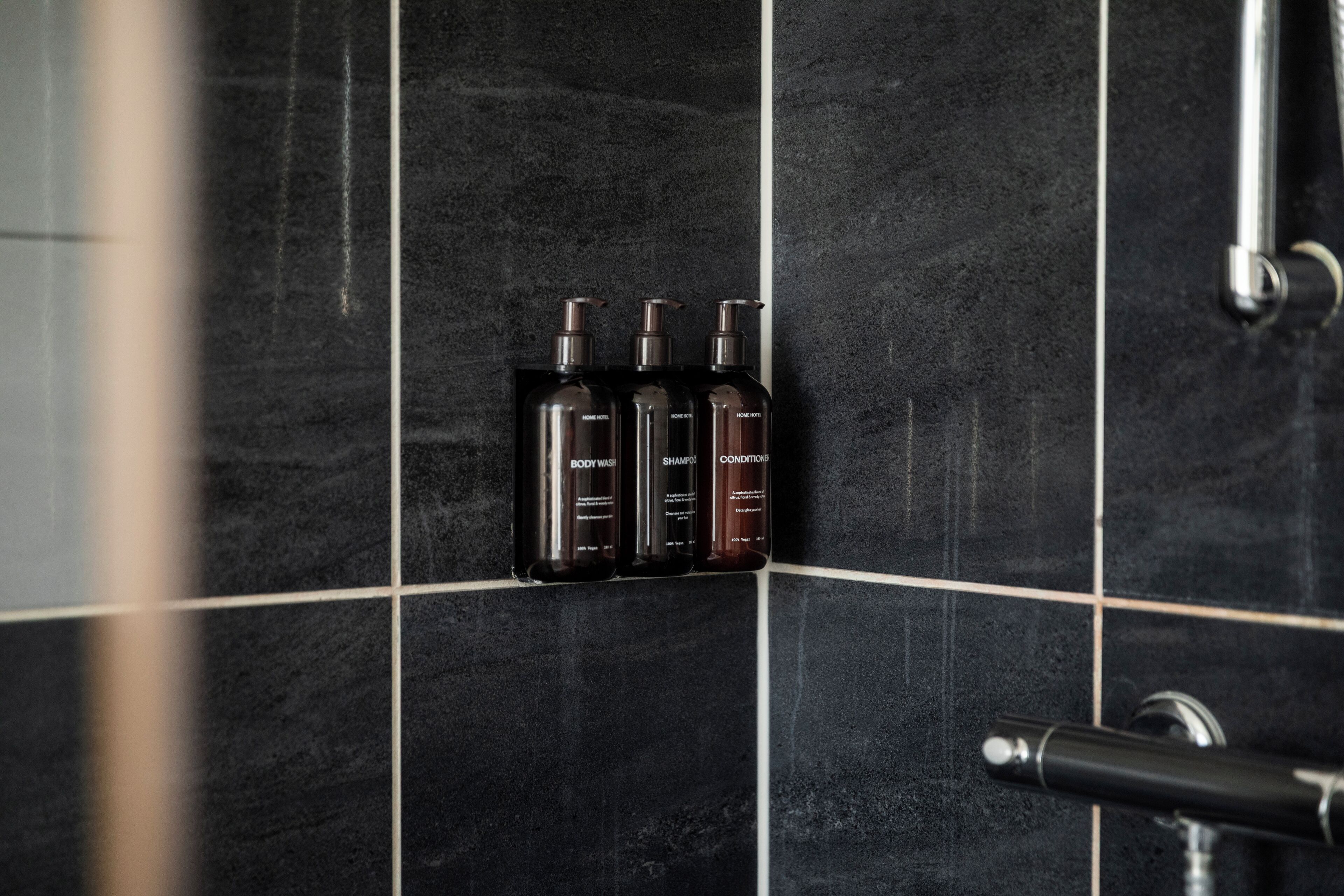 Un baño con azulejos negros y tres botellas de artículos de aseo en un estante.