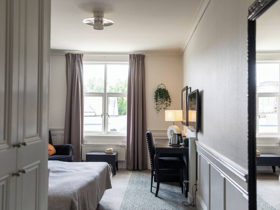 A hotel room with a bed, a desk, a chair, a lamp, and a window with curtains.