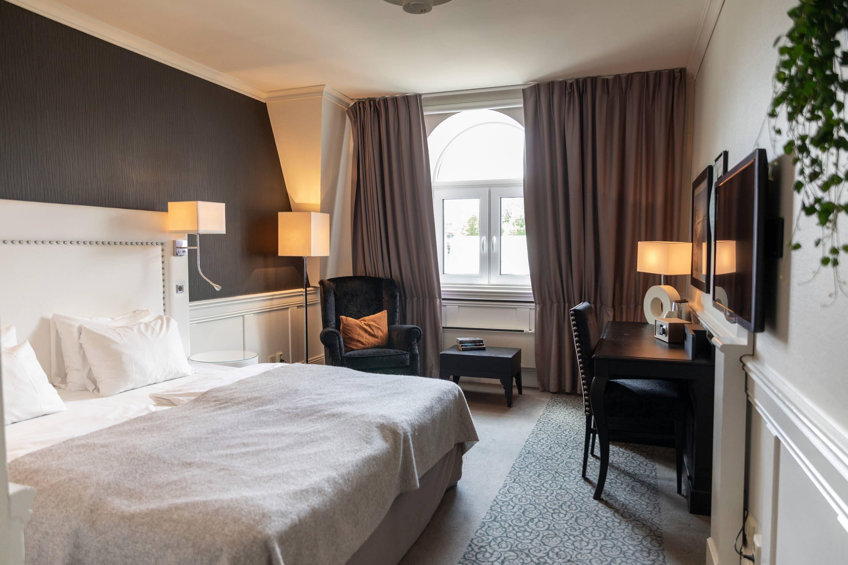 A hotel room with a large bed, a desk with a chair, a TV, and a window with curtains.