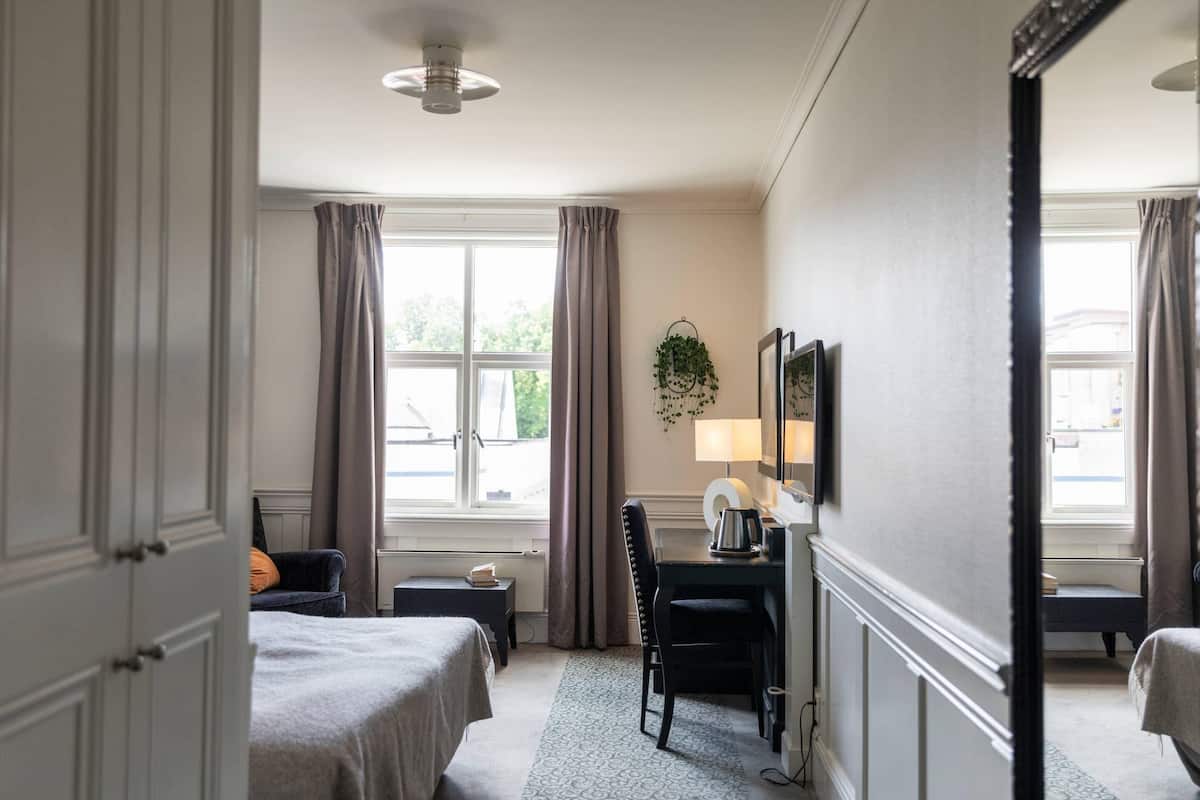 A hotel room with two beds, a desk with a lamp, a chair, a window with curtains, and a plant on the wall.