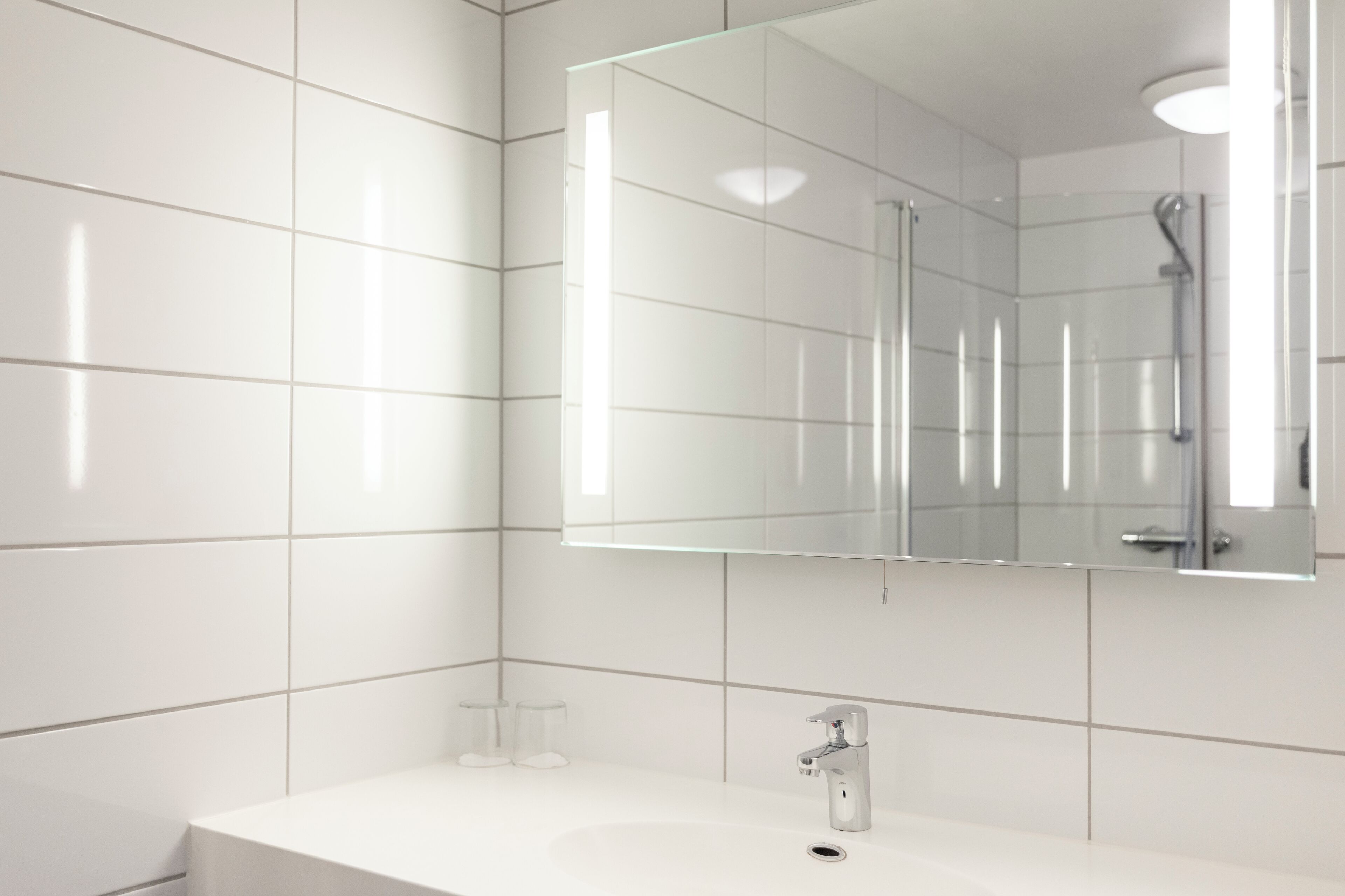 Een moderne badkamer met witte tegels, een grote spiegel, een wastafel en een douche.