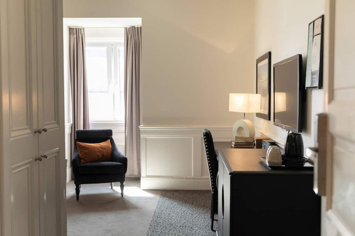 A hotel room with a dark armchair, a desk with a lamp, and a large window with curtains.