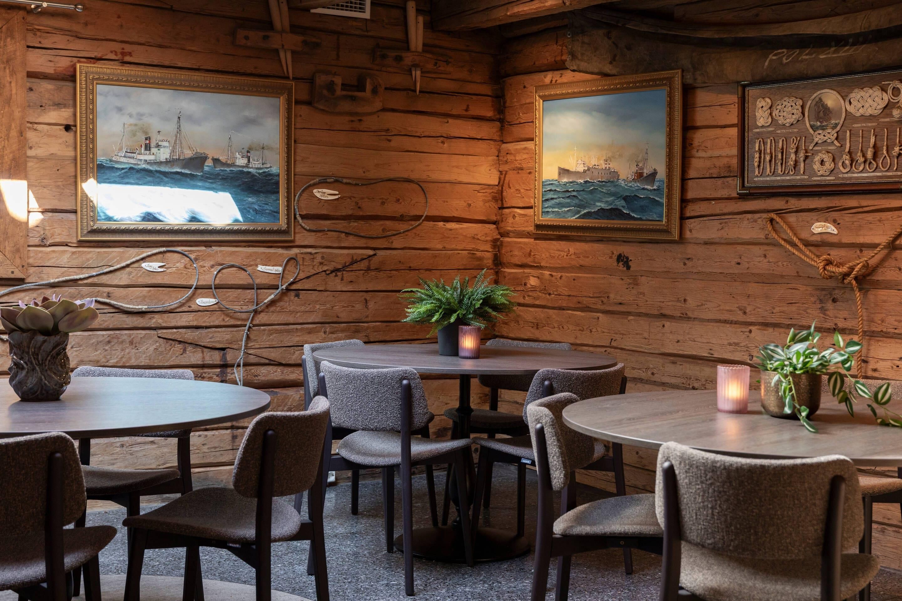 A cozy wooden cabin interior with round tables and chairs, framed maritime paintings, and a potted plant.