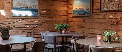 A cozy wooden cabin interior with round tables and chairs, framed maritime paintings, and a potted plant.