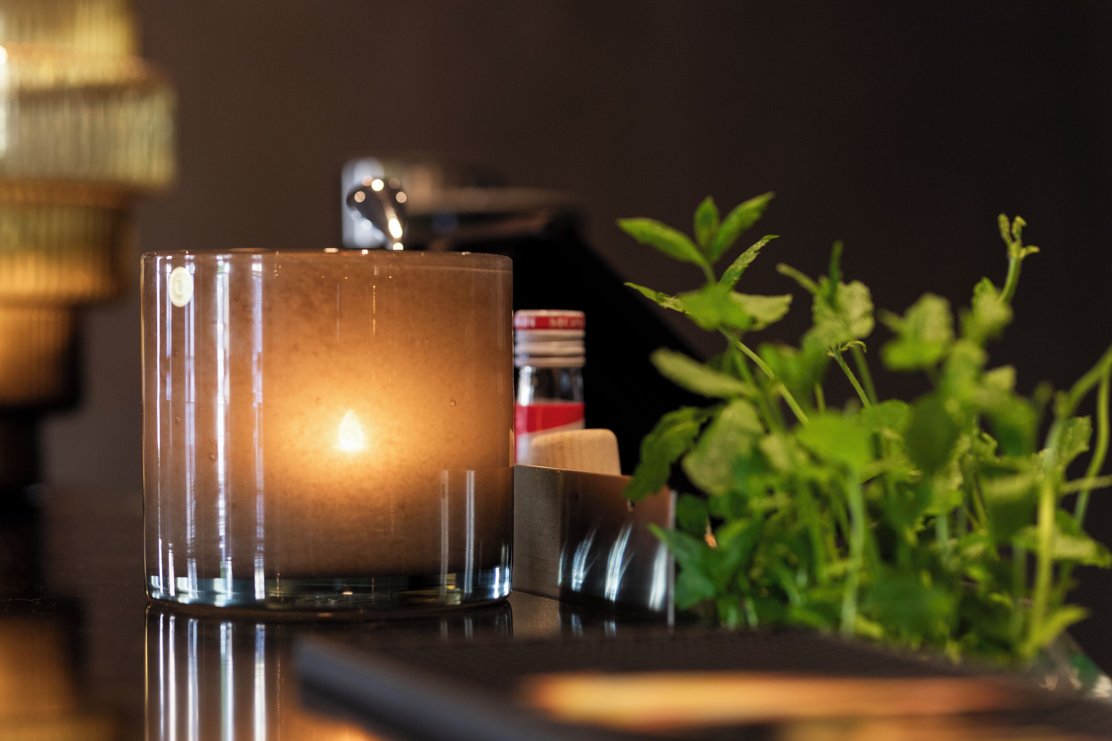 Una vela en un recipiente de vidrio sobre una superficie reflectante, con una planta y un libro al fondo.