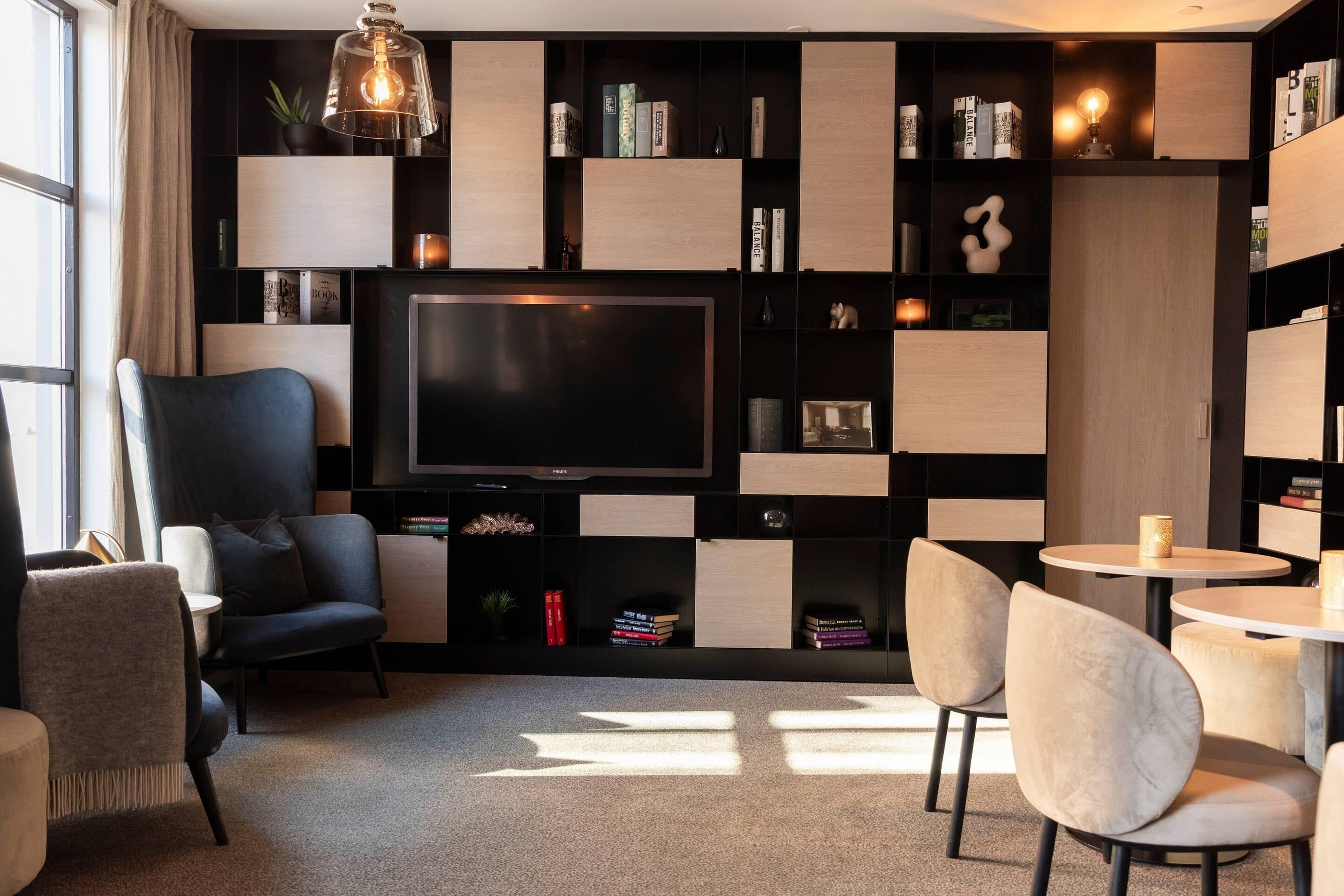 Una sala moderna con un televisor de pantalla plana grande, una zona de estar cómoda con sofá y sillones, una mesita redonda pequeña y una estantería llena de libros y objetos decorativos.