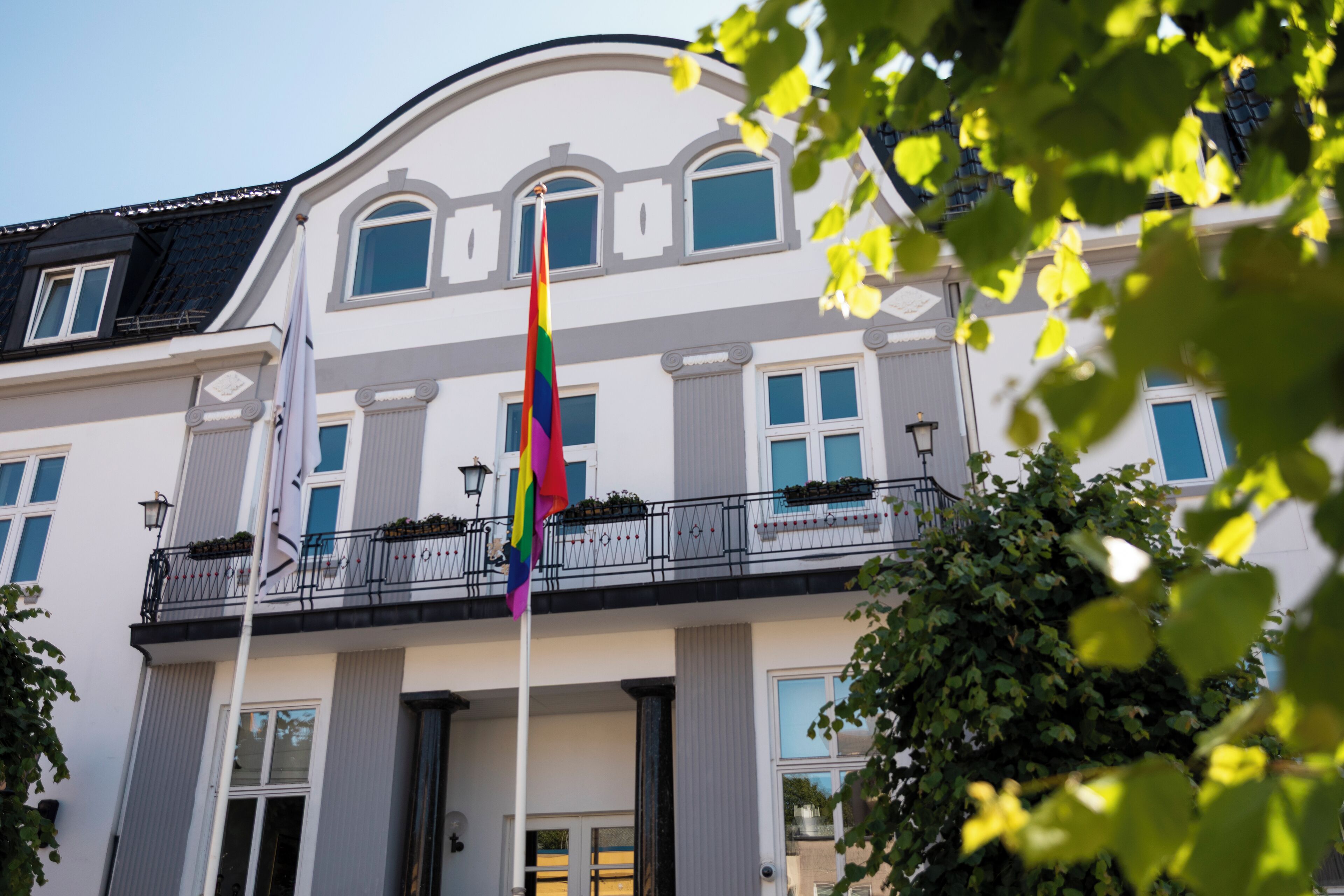 Un edificio con fachada blanca, múltiples ventanas e izadora con una bandera arcoíris.