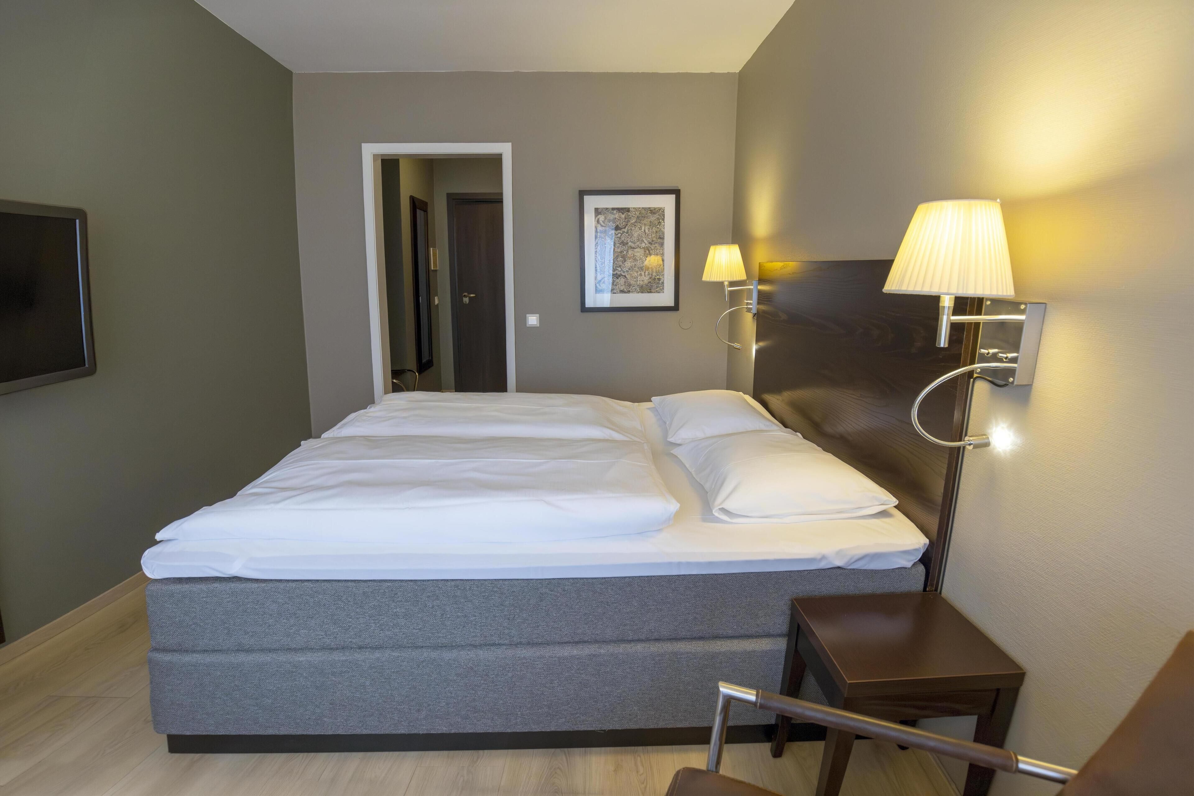 A hotel room with a large bed, a bedside table with a lamp, a small table, and a TV mounted on the wall.
