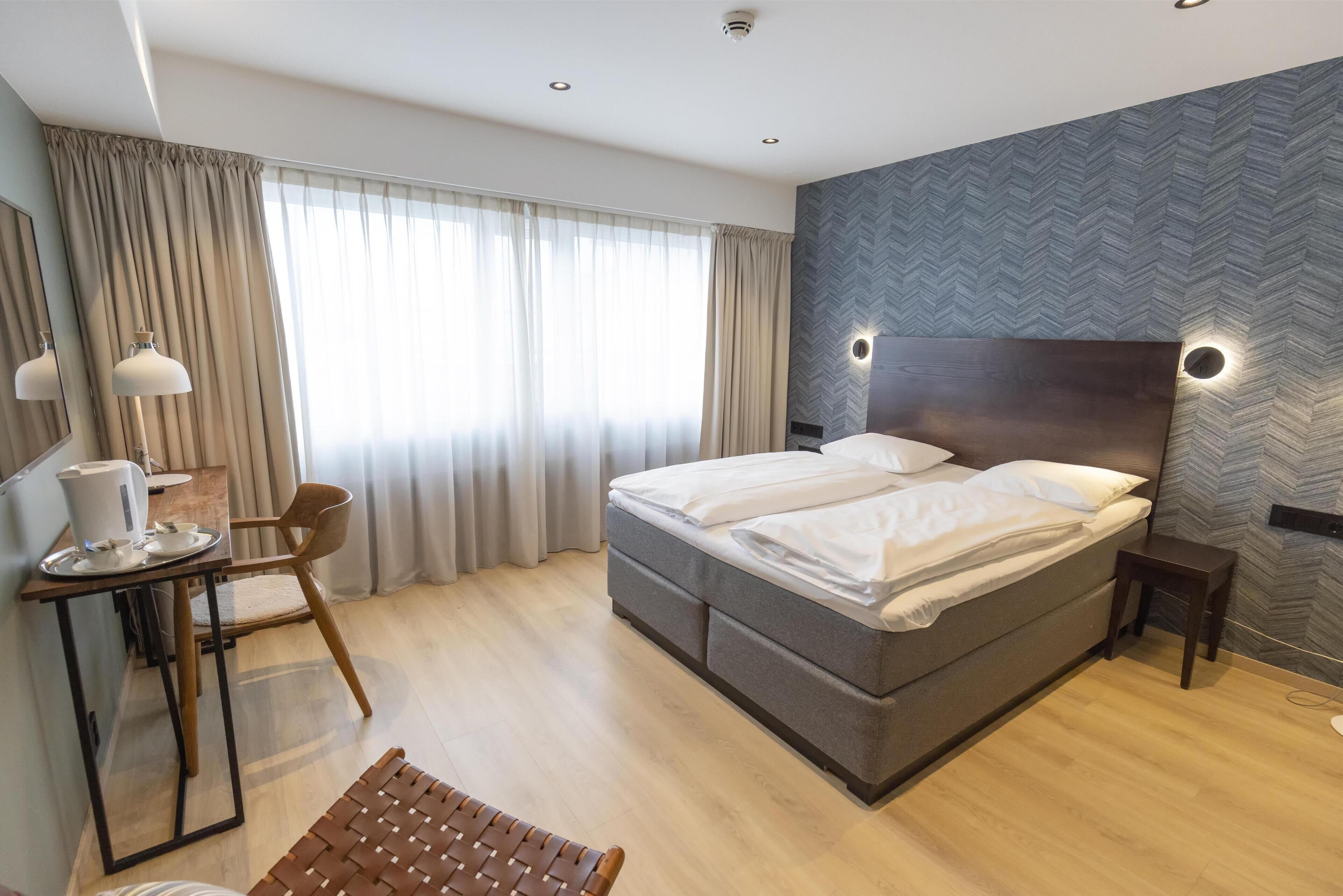 A modern hotel room with a large bed, a desk with a lamp, a chair, and a small table with a kettle.