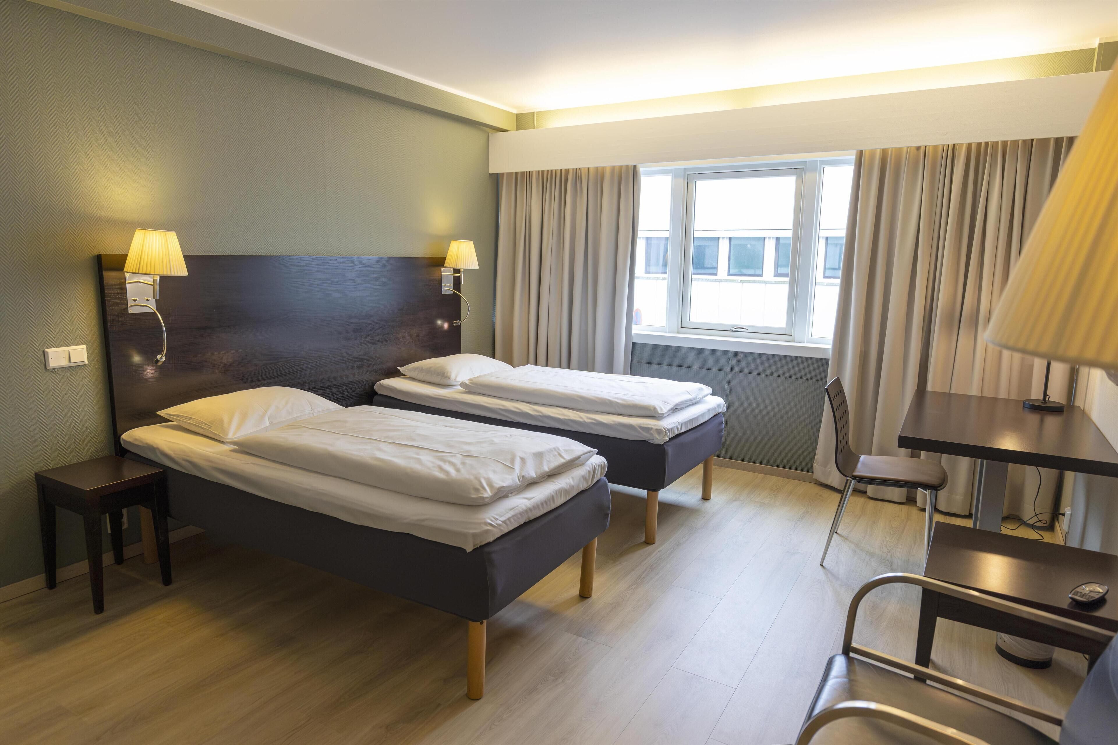 A hotel room with two single beds, a desk, a chair, a lamp, and a window with curtains.