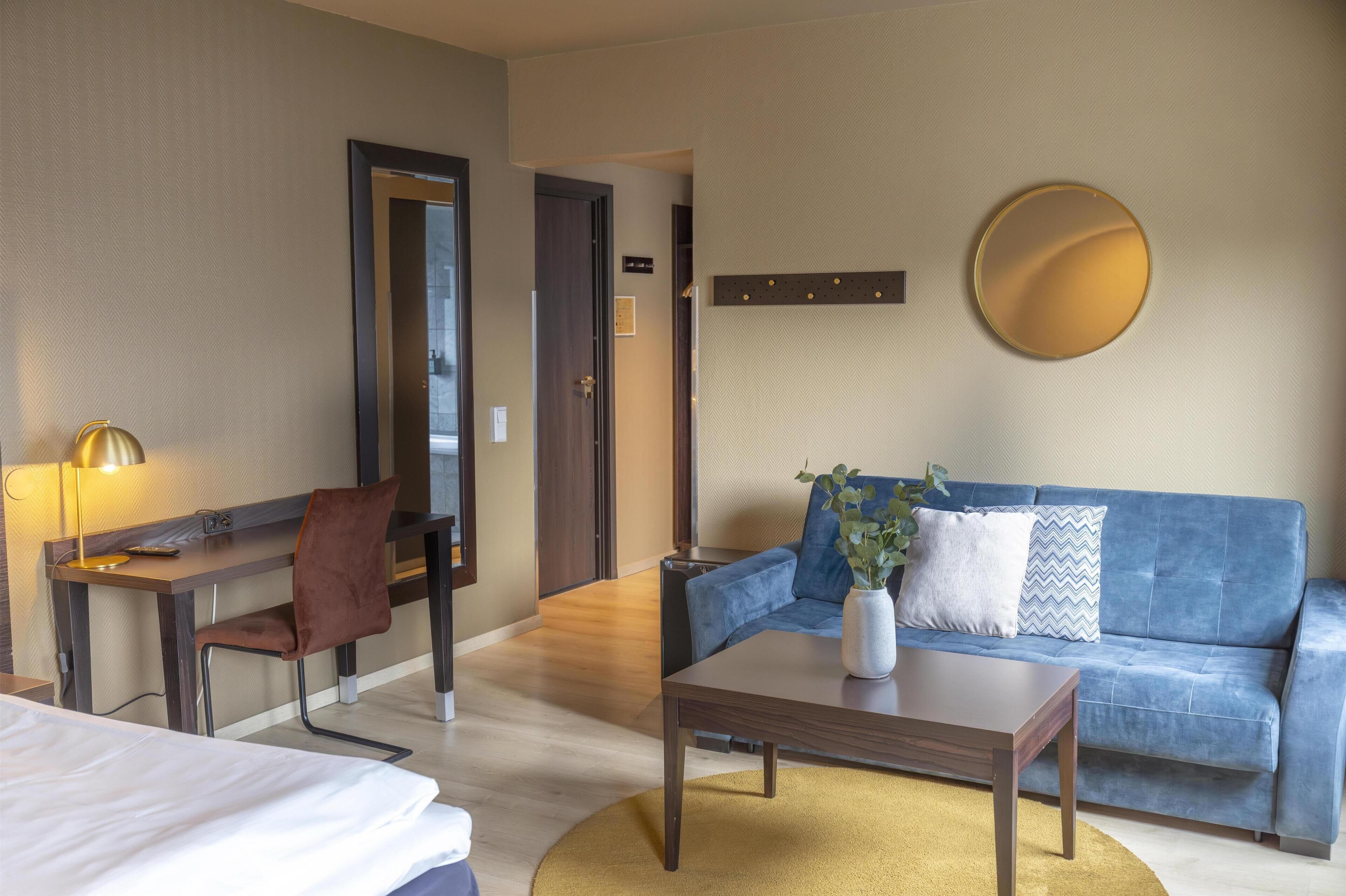 Una habitación de hotel con cama, escritorio y silla, un sofá azul, una mesa de centro y un espejo redondo en la pared.