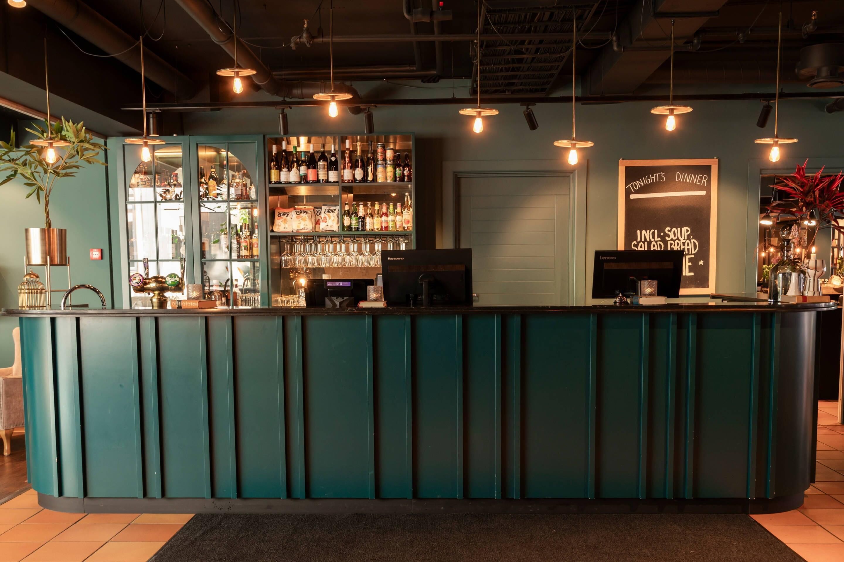 Una zona de bar con mostrador verde azulado, estantes con botellas, un menú en la pizarra y luces colgantes.