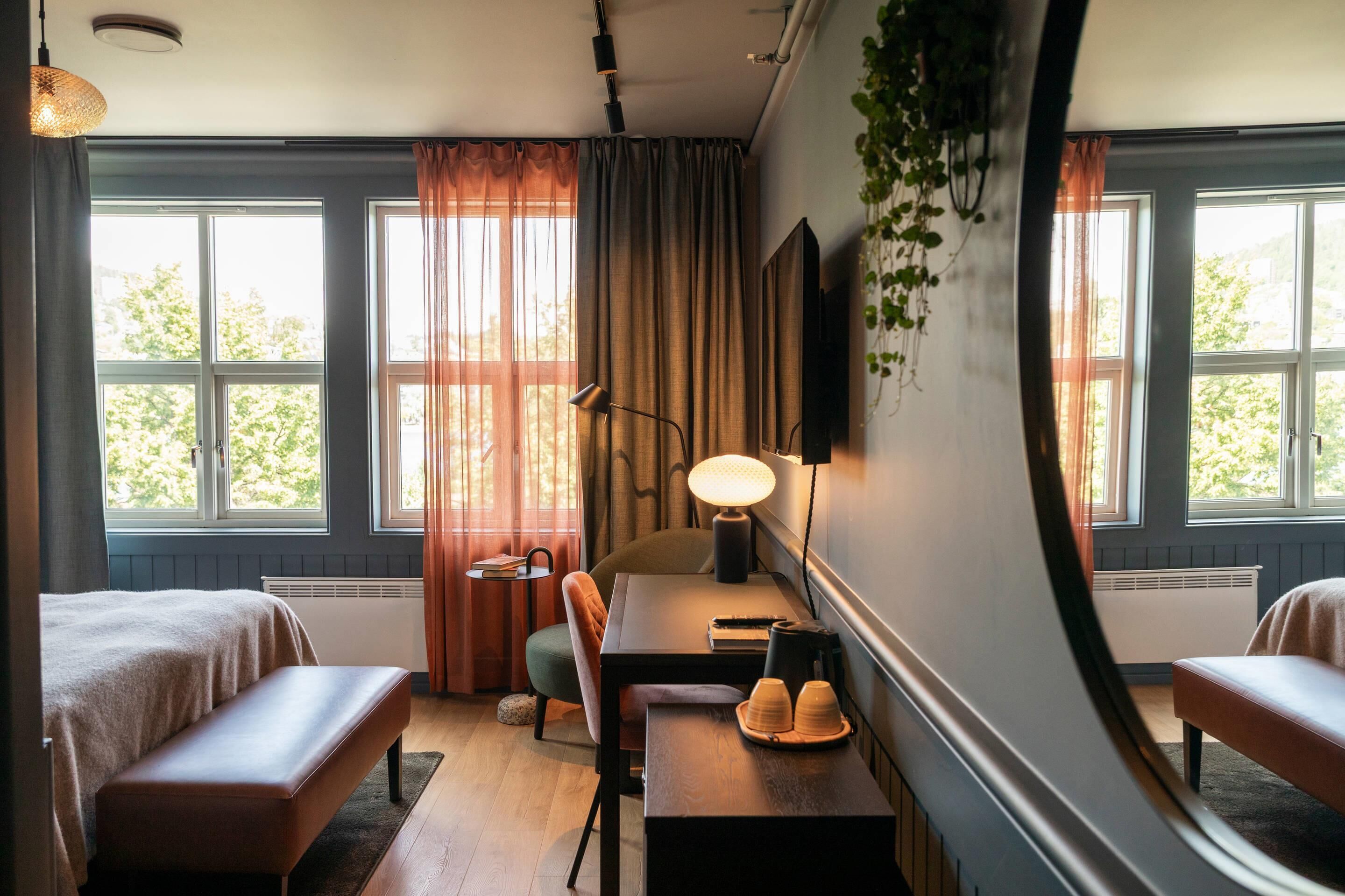 Habitación de hotel con dos camas, un escritorio con lámpara, una silla y vistas a árboles a través de las ventanas.