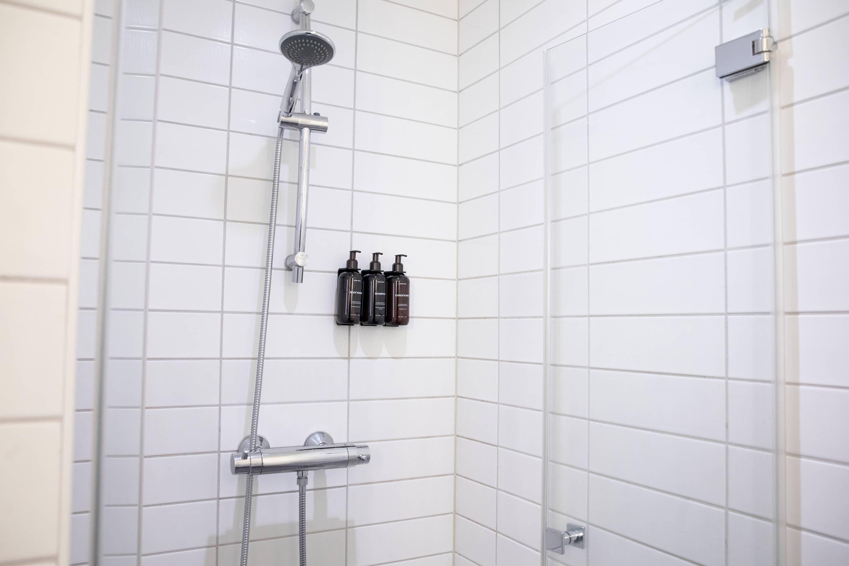 A shower with a handheld showerhead and a handheld hose, a wall-mounted control panel, and three bottles of shampoo on the wall.
