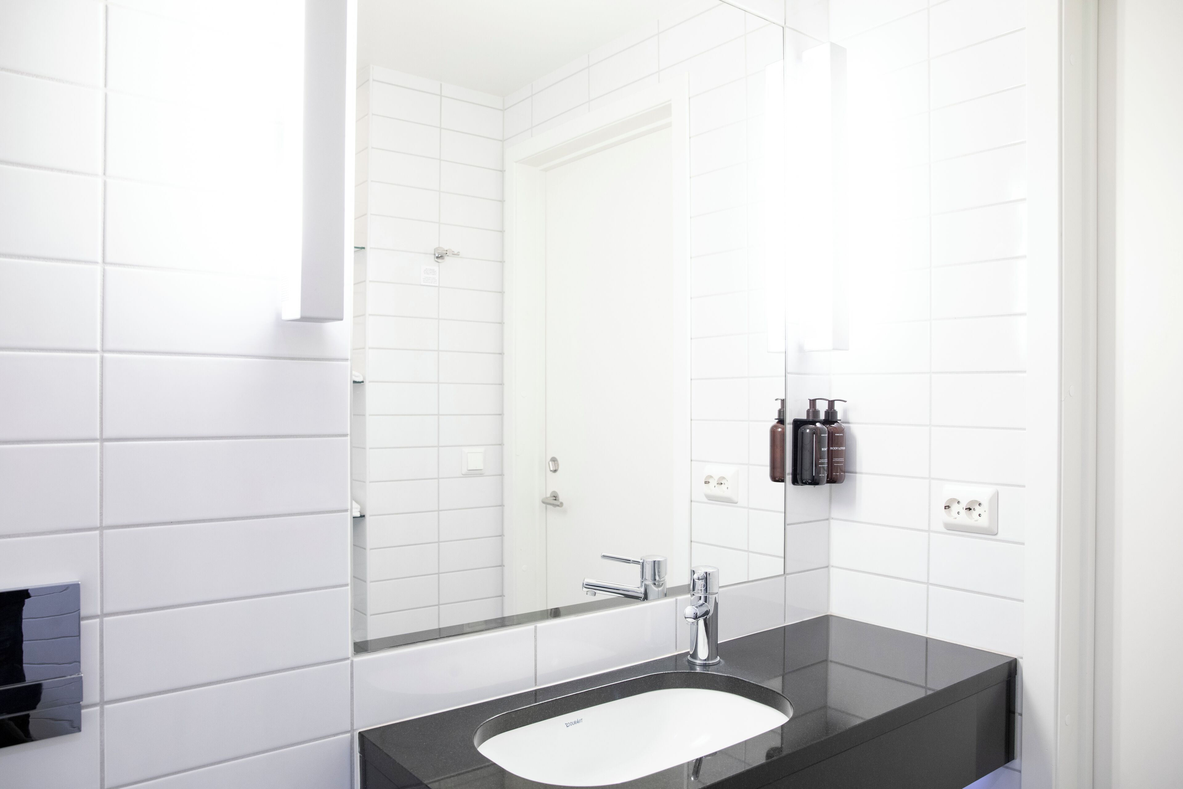 A modern bathroom with white tiles, a black sink, and a mirror.