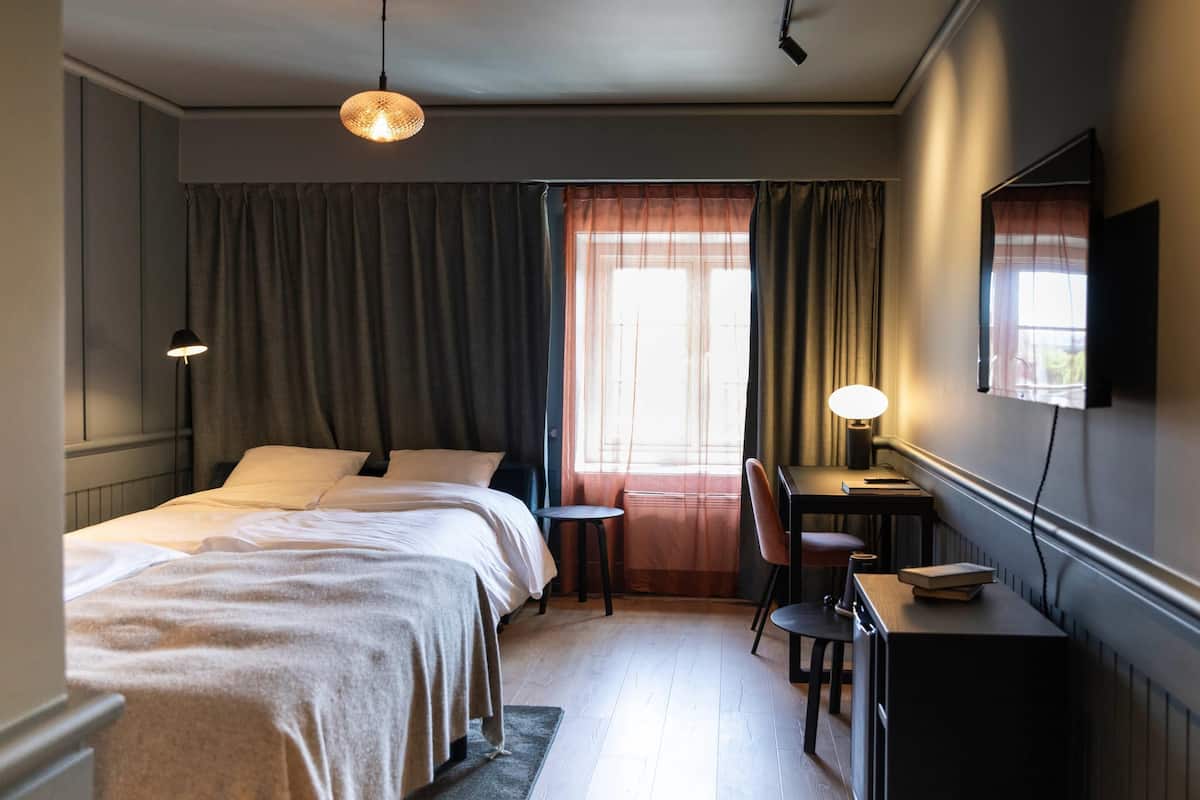A hotel room with a bed, desk, chair, lamp, and a window with curtains.