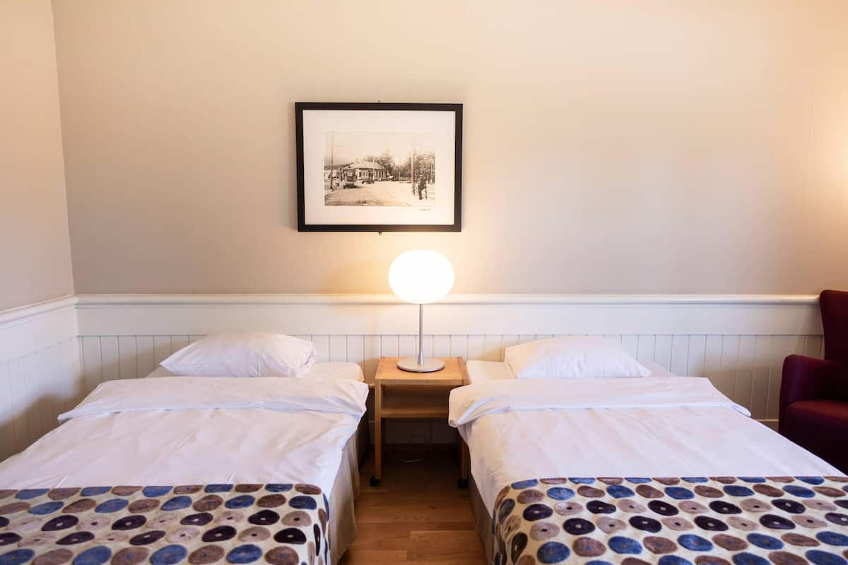 A hotel room with two beds, a nightstand with a lamp, a framed picture on the wall, and a red chair.